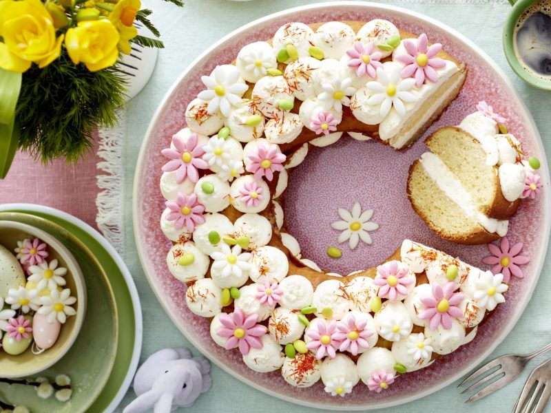 Frühlingskranz auf einem rosa Teller auf einem grünen Geschirrtuch. Rechts Gabeln und links auf dem Foto kleine Teller, Oster-Deko und Blumen. Rechts oben auf dem Foto eine Tasse Kaffee.