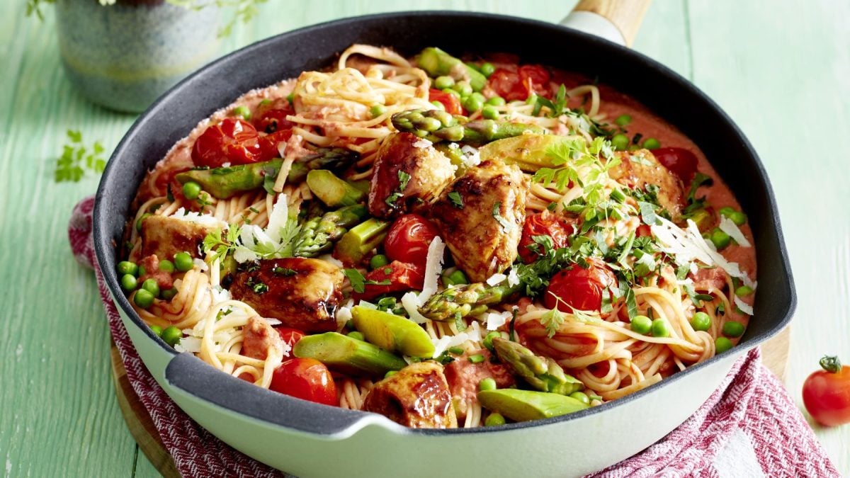 Hähnchen-Spargel-Pfanne in Tomatenrahm in einer schwarz-weißen Pfanne auf grünem Hintergrund. Rechts unten auf dem Foto ein rot-weiß-kariertes Geschirrtuch.