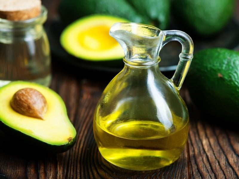 Frontal: Eine kleine Karaffe aus Glas mit einer hellen, leicht öligen Flüssigkeit. Drum herum liegen zwei Avocado-Hälften. Die eine hat einen Kern und die andere nicht.