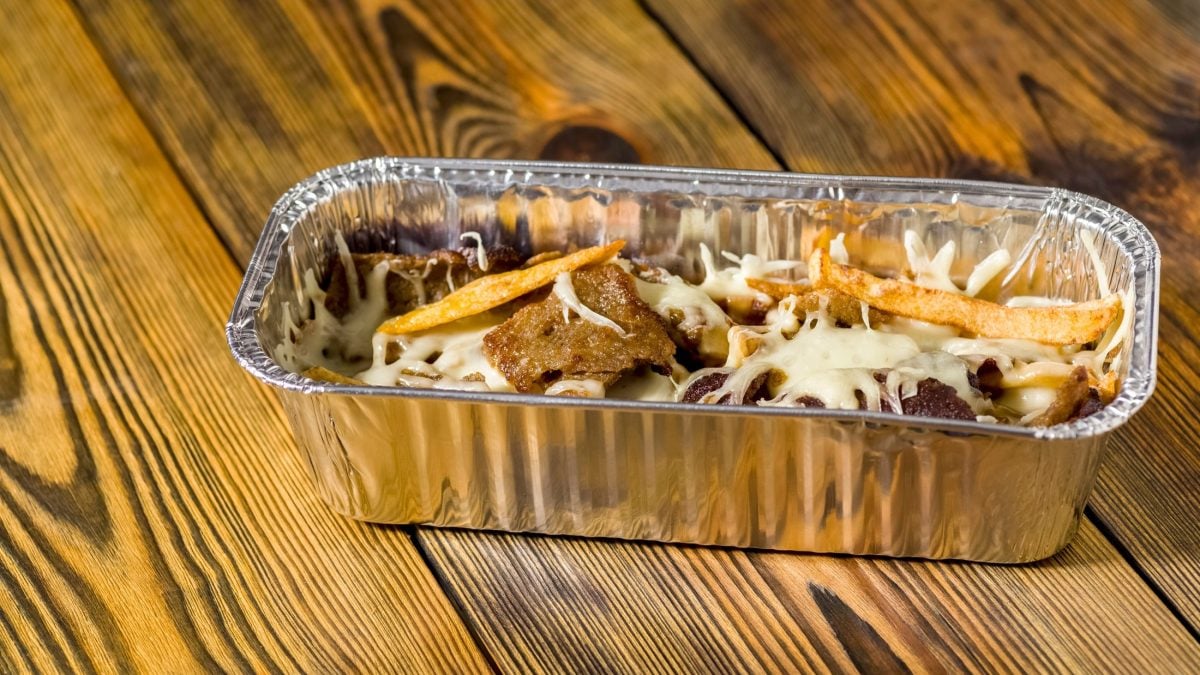 Draufsicht, schräg: Holzboden, eine Aluschale in der dunkel gebratenen Fleischstücke mit einer hellen Sauce und Zwiebelringen sind.