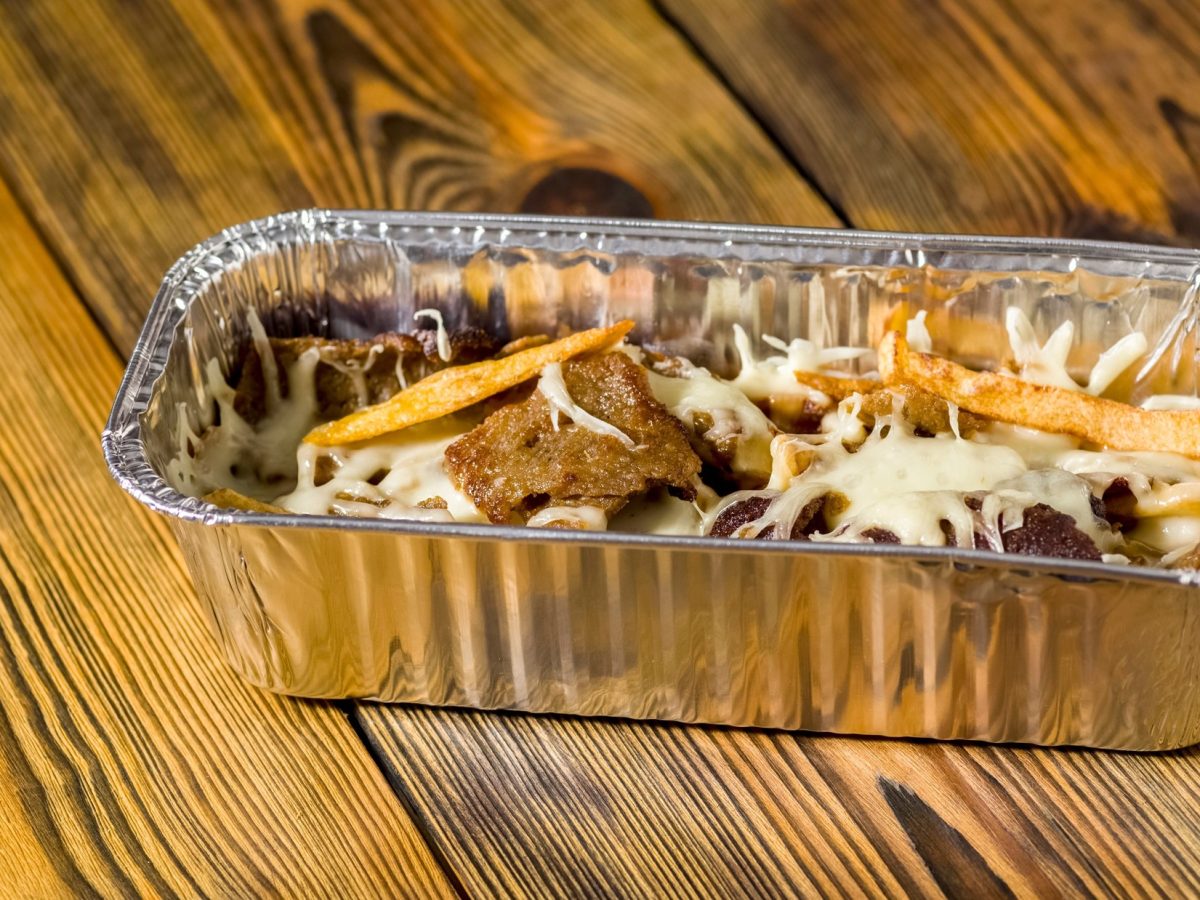Draufsicht, schräg: Holzboden, eine Aluschale in der dunkel gebratenen Fleischstücke mit einer hellen Sauce und Zwiebelringen sind.