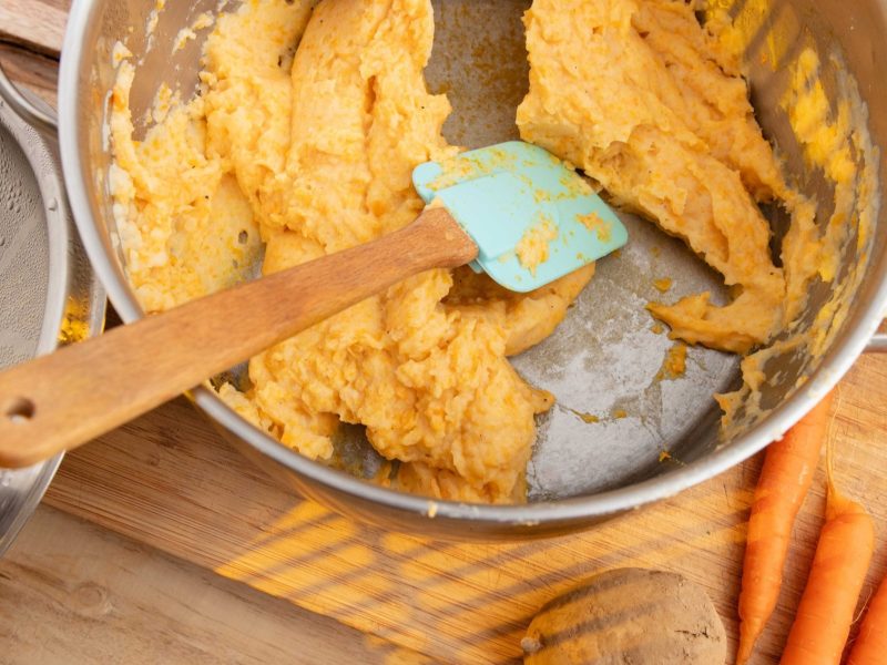 Draufsicht schräg / Kartoffel-Möhren-Stampf mit Curry n einem Topf. Daneben liegen ein Topfdeckeln, frische Möhren und eine Kartoffel.