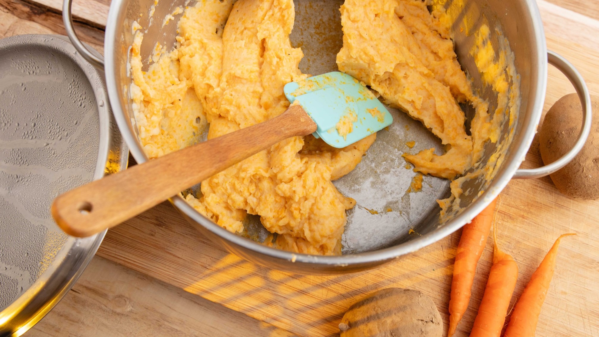 Draufsicht schräg / Kartoffel-Möhren-Stampf mit Curry n einem Topf. Daneben liegen ein Topfdeckeln, frische Möhren und eine Kartoffel.