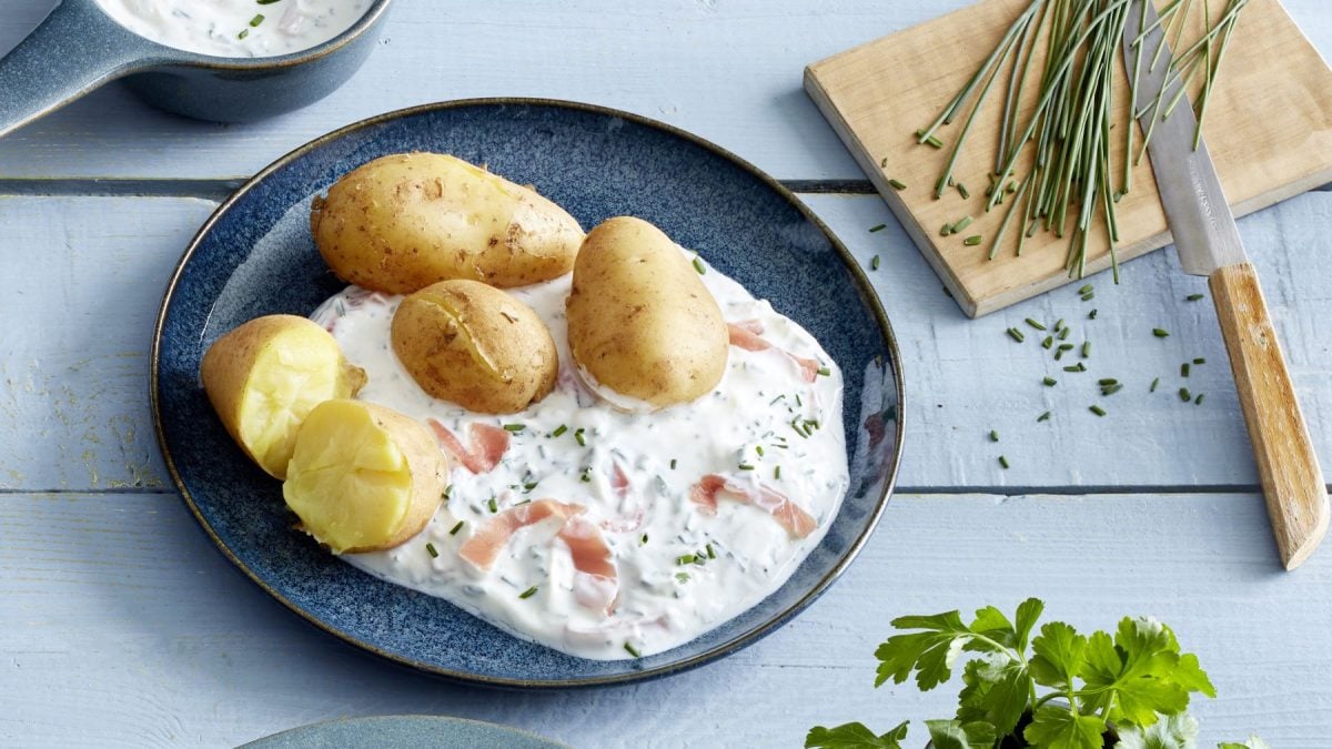Kartoffeln mit Schnittlauch auf einem blauen Teller auf hellblauem Hintergrund. Rechts auf dem Bild ein Schneidebrett mit Schnittlauch und unten rechts Petersilie.