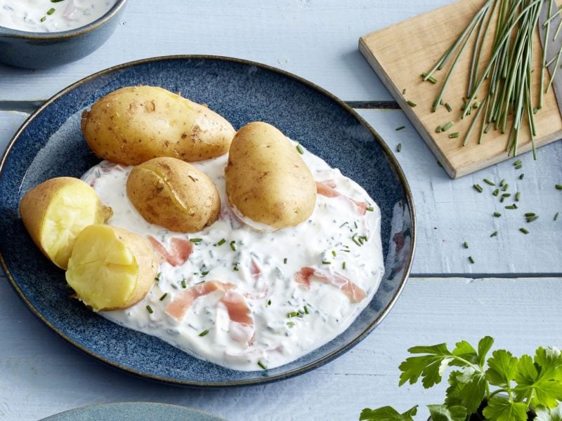 Kartoffeln mit Schnittlauch auf einem blauen Teller auf hellblauem Hintergrund. Rechts auf dem Bild ein Schneidebrett mit Schnittlauch und unten rechts Petersilie.