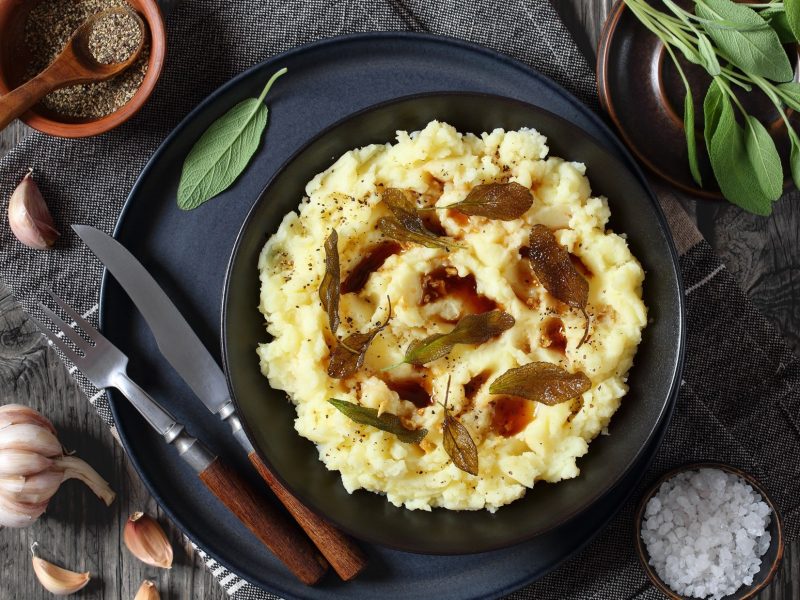 Kartoffelpüree mit gebratenen Salbeiblättern in schwarzer Schüssel auf rustikal dunklem Holztisch mit Besteck, frischem Salbei, Salz und Pfeffer, horizontale Ansicht von oben, flache Lage
