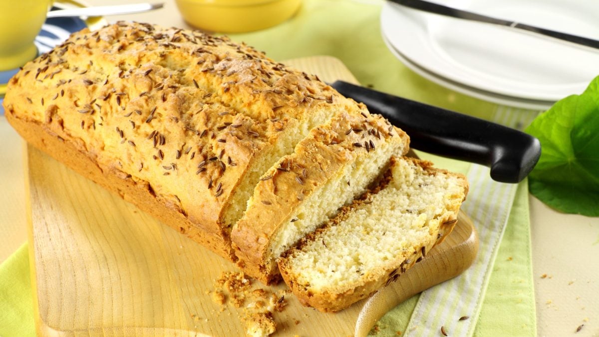 Frontal: Ein kastenförmiger Kuchen auf einem Holzbrett. Daneben ein Küchenmesser und ein runder, heller Teller.