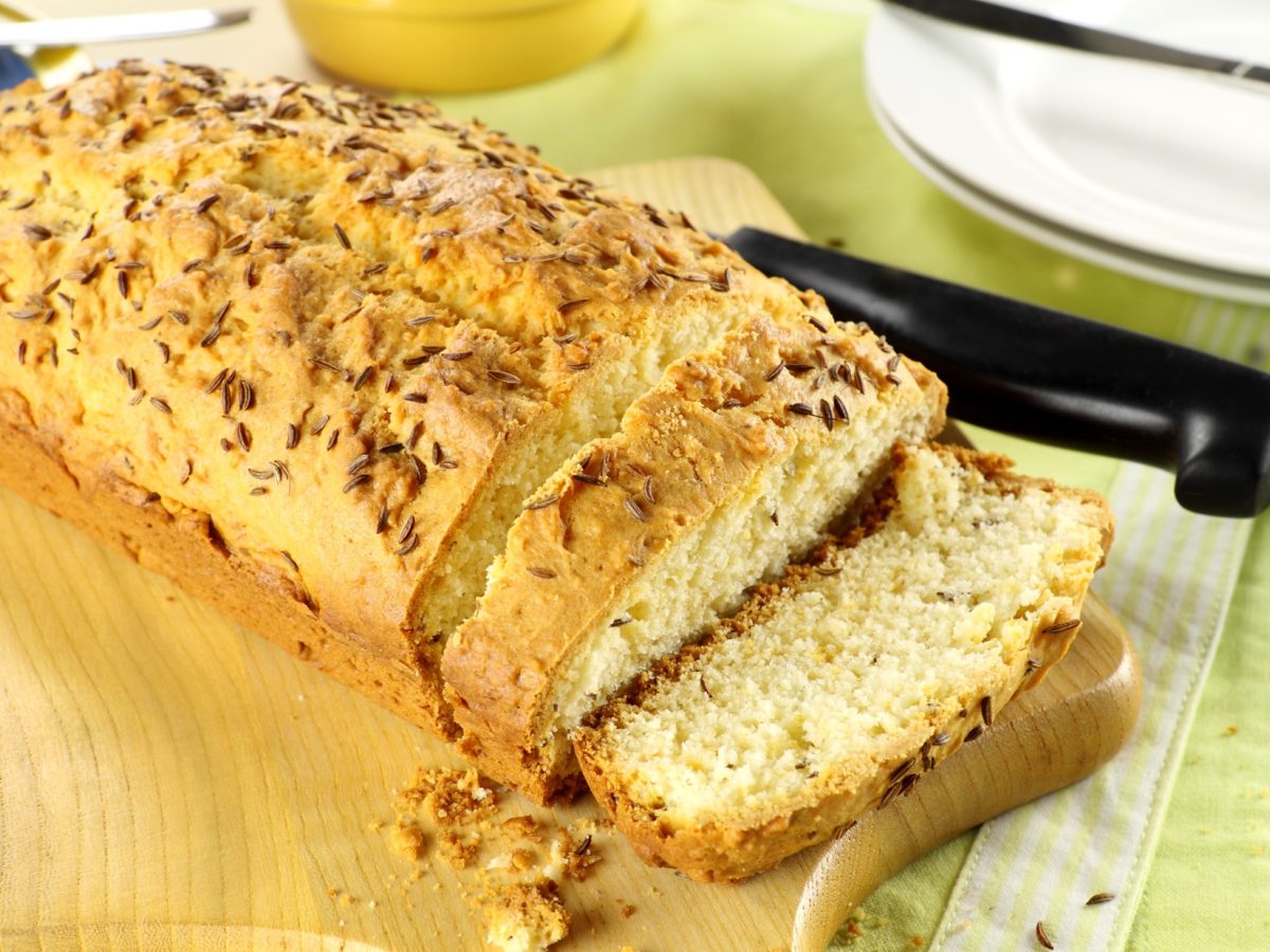 Frontal: Ein kastenförmiger Kuchen auf einem Holzbrett. Daneben ein Küchenmesser und ein runder, heller Teller.