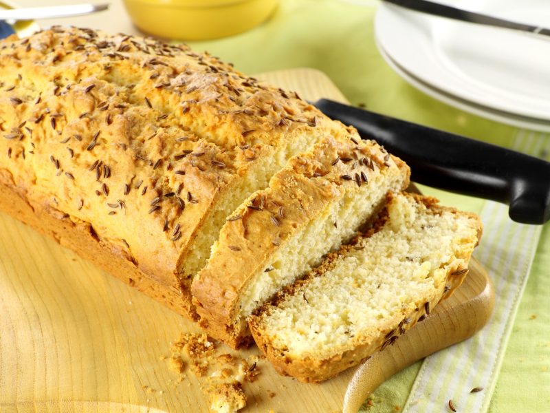 Frontal: Ein kastenförmiger Kuchen auf einem Holzbrett. Daneben ein Küchenmesser und ein runder, heller Teller.
