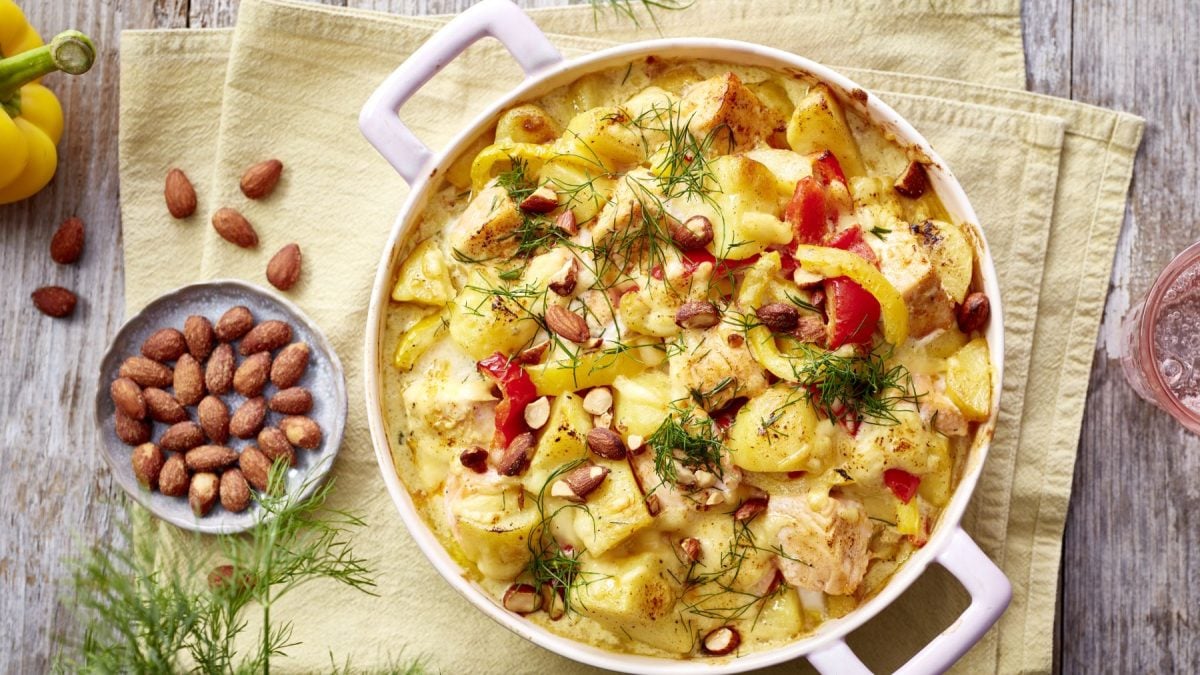 Lachs-Kartoffel-Auflauf auf einem gelben Geschirrtuch auf hölzernem Hintergrund. Links ein Schälchen mit Mandeln und Dill.