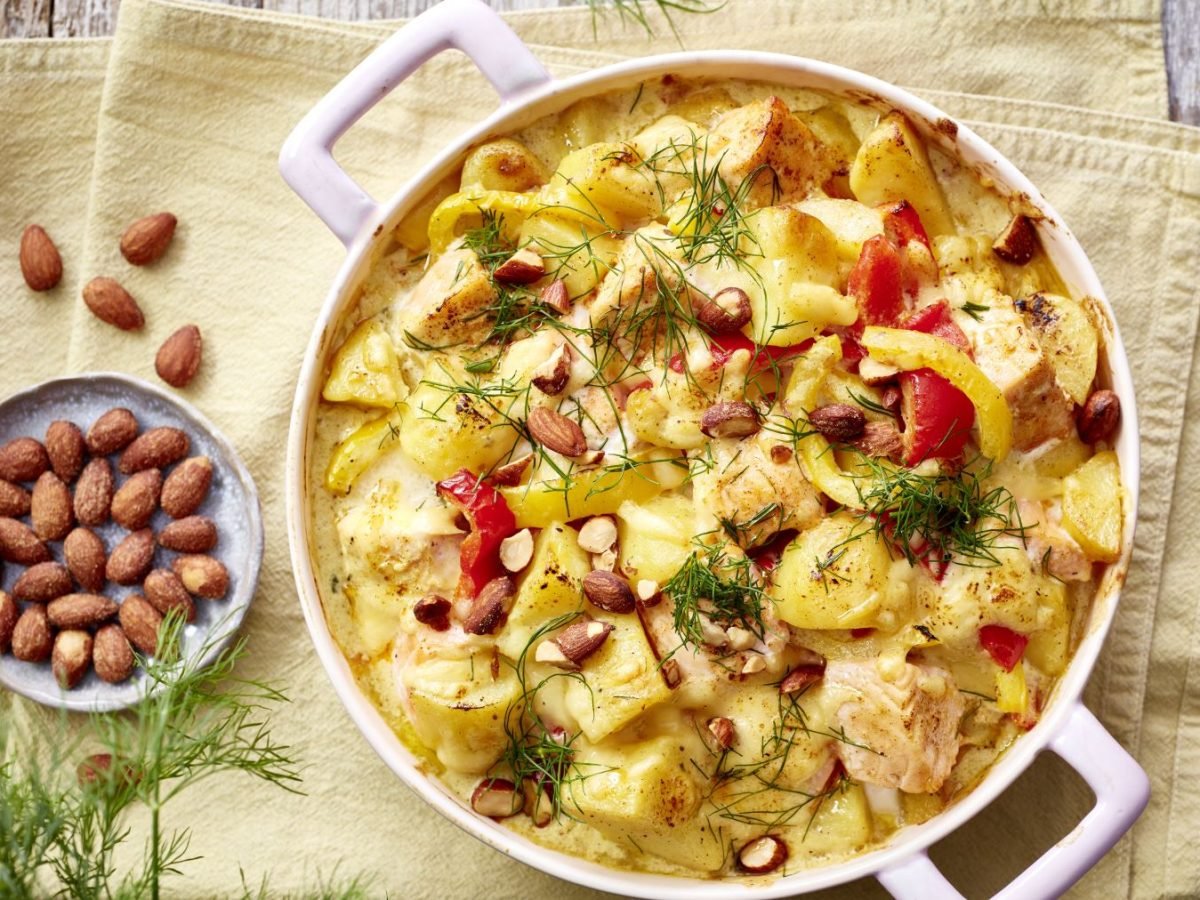 Lachs-Kartoffel-Auflauf auf einem gelben Geschirrtuch auf hölzernem Hintergrund. Links ein Schälchen mit Mandeln und Dill.