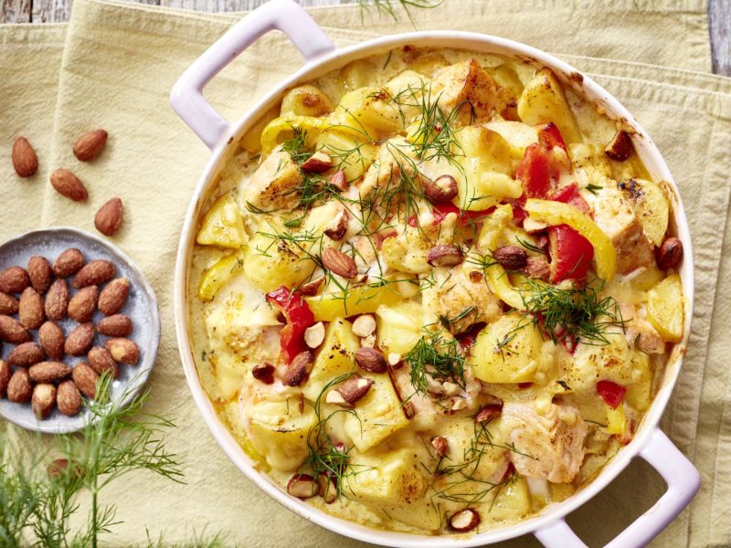 Lachs-Kartoffel-Auflauf auf einem gelben Geschirrtuch auf hölzernem Hintergrund. Links ein Schälchen mit Mandeln und Dill.