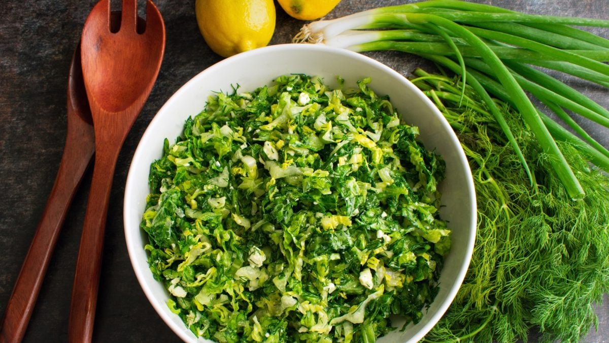 Draufsicht: Eine runde Schüssel Maroulosalata auf einem dunklem Untergrund. Daneben: Holzlöffel, Zitronen und Frühlingszwiebeln.