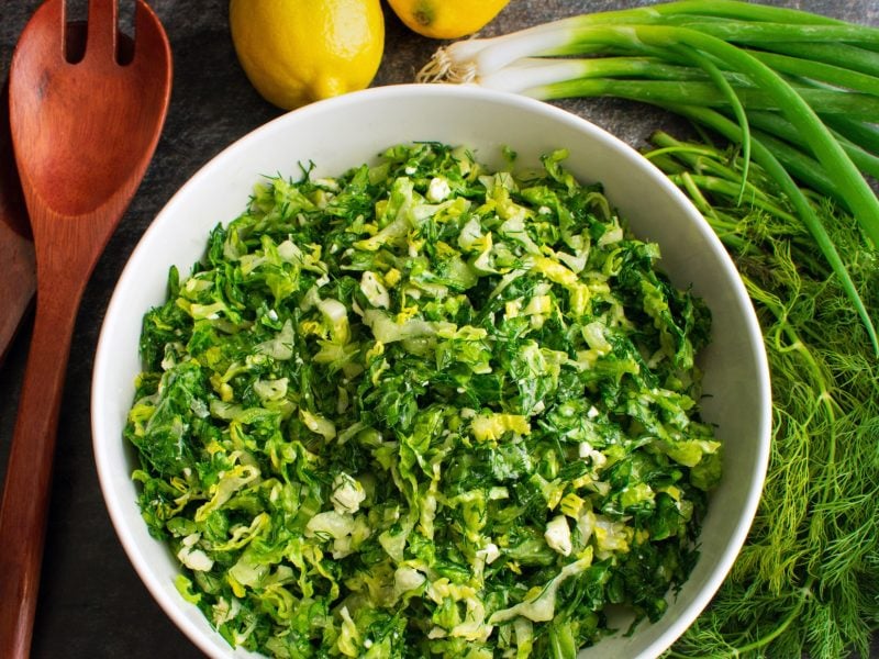 Draufsicht: Eine runde Schüssel Maroulosalata auf einem dunklem Untergrund. Daneben: Holzlöffel, Zitronen und Frühlingszwiebeln.