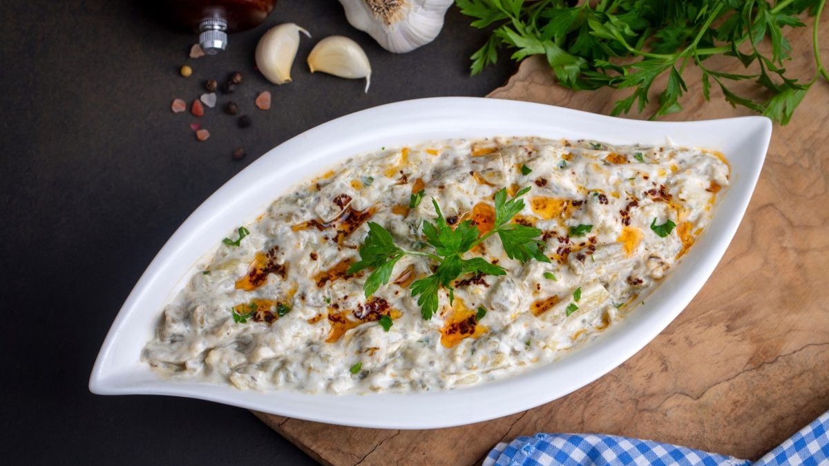 Draufsicht: Eine geschwungene Schale aus Porzellan. In der Schale ist er orientalische Dip Mutabbal. Daneben liegen frische Knoblauchzehen.