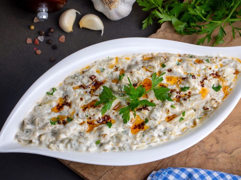 Draufsicht: Eine geschwungene Schale aus Porzellan. In der Schale ist er orientalische Dip Mutabbal. Daneben liegen frische Knoblauchzehen.