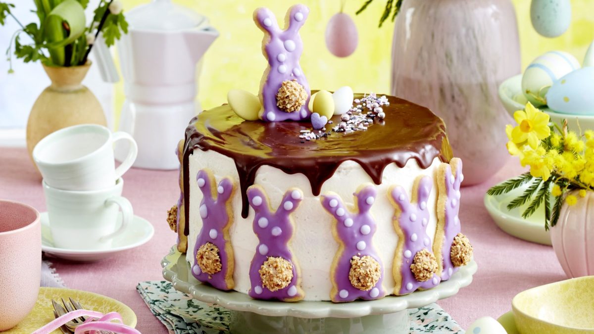 Osterhasen-Quark-Torte auf einem Tortenteller auf rosa Hintergrund. Hinten auf dem Foto Geschirr, Osterdeko und Blumen verteilt.