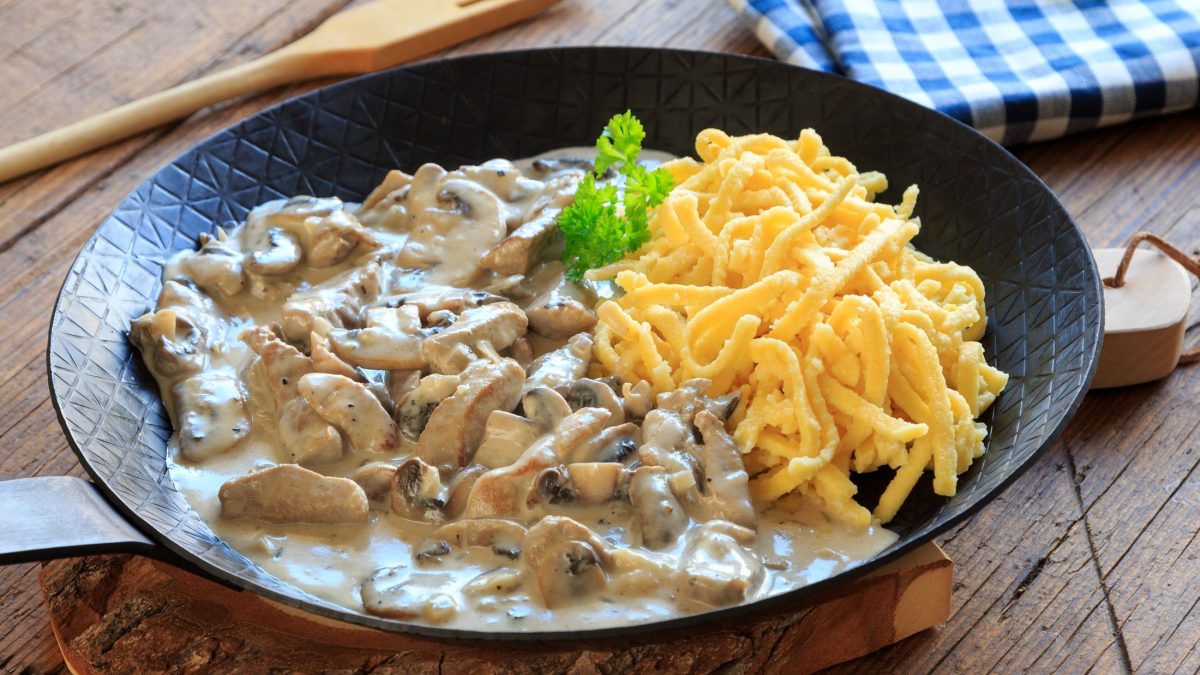 Draufsicht, schräg: Putengeschnetzeltes mit Nudeln in einer gusseisernen Pfanne mit Stiel auf einem Untergrund aus Holz.