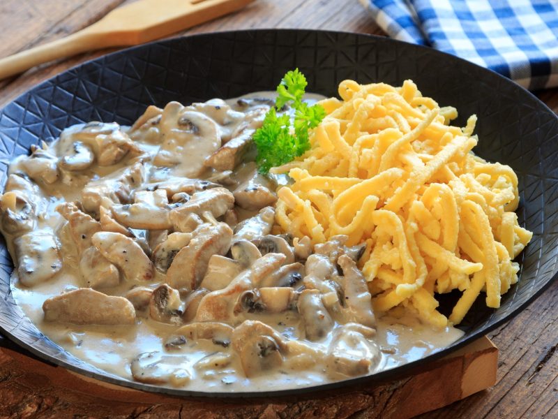 Draufsicht, schräg: Putengeschnetzeltes mit Nudeln in einer gusseisernen Pfanne mit Stiel auf einem Untergrund aus Holz.