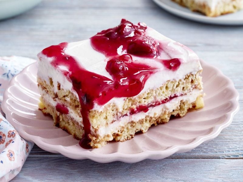 Rote-Grütze-Tiramisu-Schnitten auf einem rosa Teller vor grauem Hintergrund. Links ein Geschirrtuch mit Blumen-Muster.
