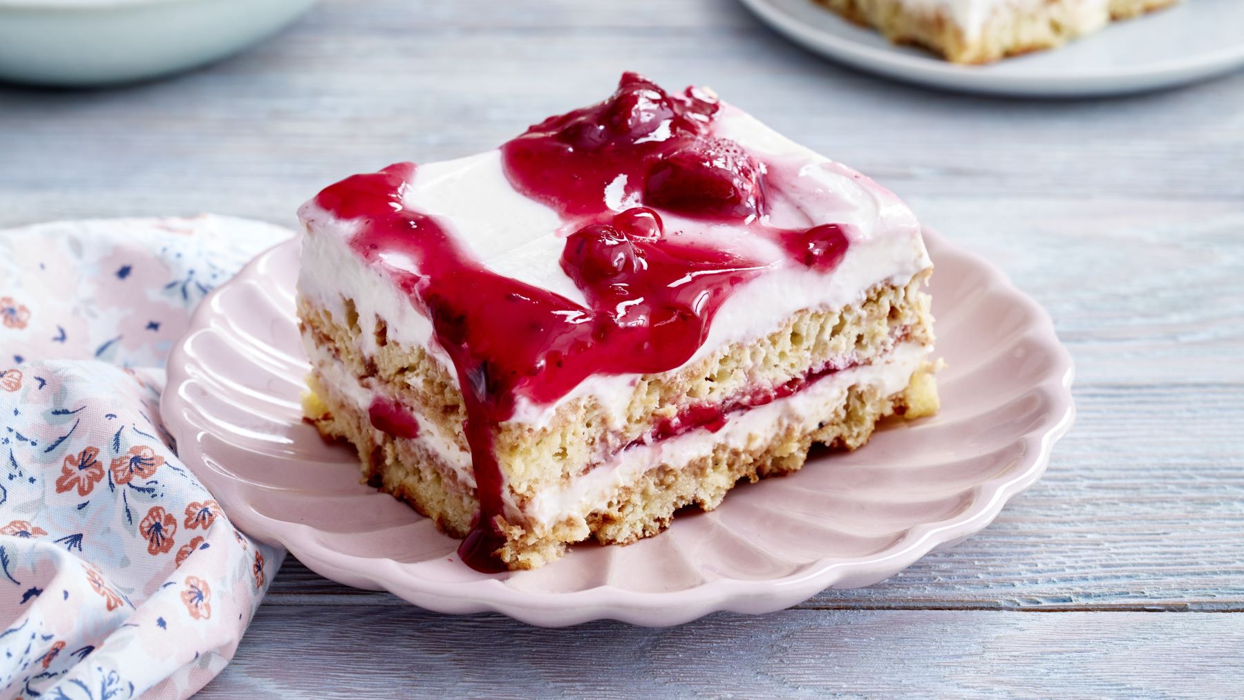 Rote-Grütze-Tiramisu-Schnitten auf einem rosa Teller vor grauem Hintergrund. Links ein Geschirrtuch mit Blumen-Muster.