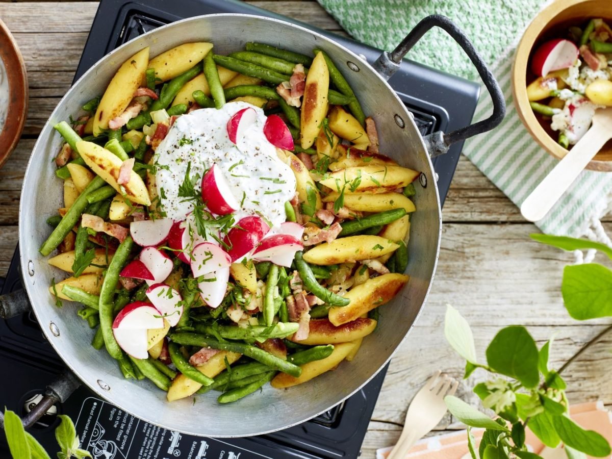Schupfnudel-Pfanne vor hölzernem Hintergrund. Links Kräuterquark, rechts ein grün-weißes Geschirrtuch und ein Schüsselchen mit einer kleinen Portion Schupfnudel-Pfanne.