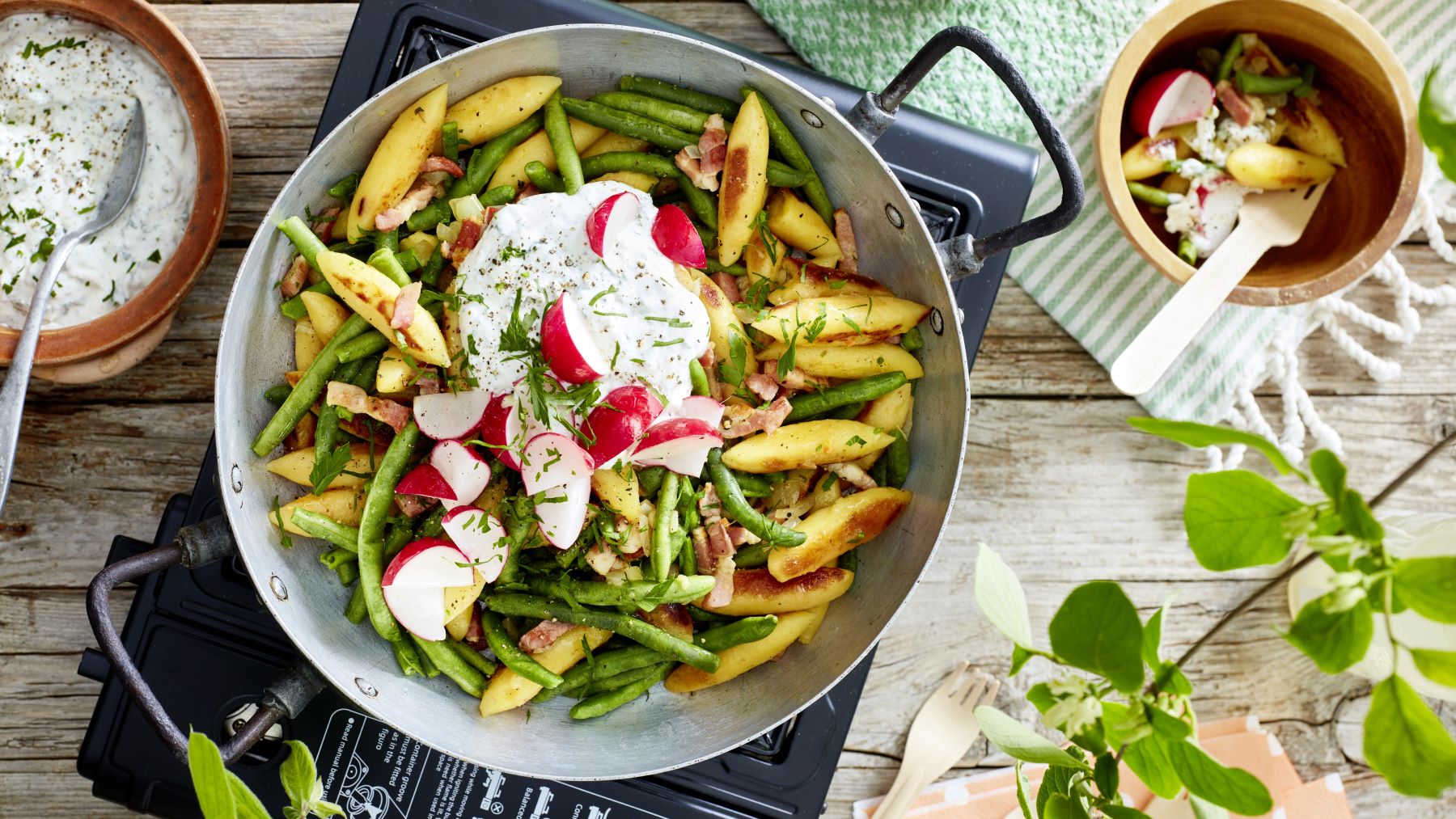 Schupfnudel-Pfanne vor hölzernem Hintergrund. Links Kräuterquark, rechts ein grün-weißes Geschirrtuch und ein Schüsselchen mit einer kleinen Portion Schupfnudel-Pfanne.