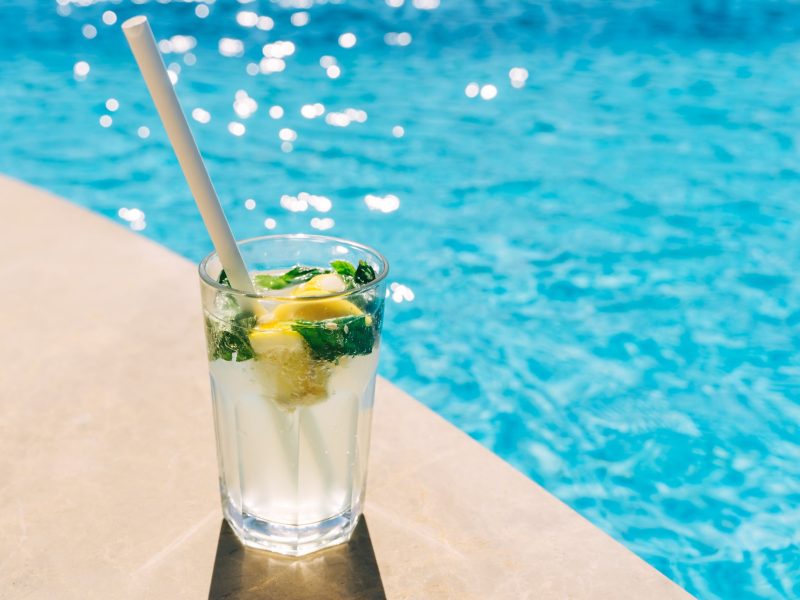 Leicht schräg von oben: Ein Glas Wasser mit Eis und frischer Zitrone in dem ein Strohhalm steckt steht am Rand eines Swimmingpools.