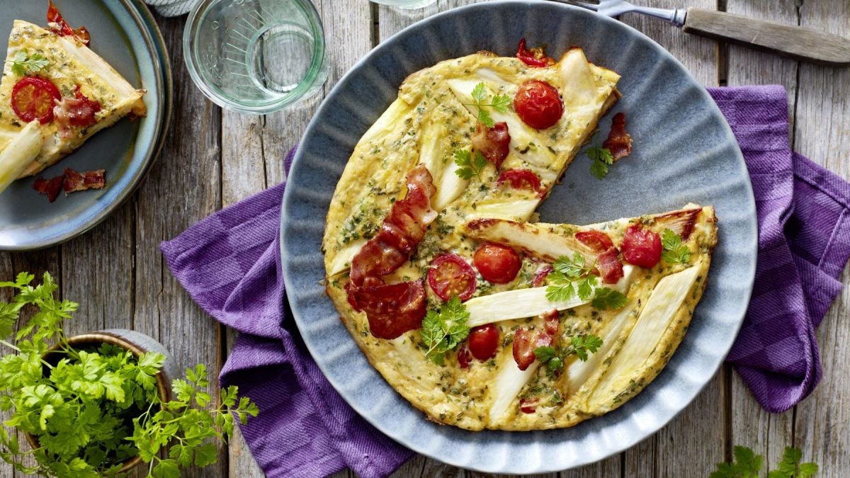 Spargel-Frittata auf einem blauen Teller auf einem lila Geschirrtuch vor hölzernem Hintergrund. Links davon Petersilie und ein Teller mit einem Stück Frittata.