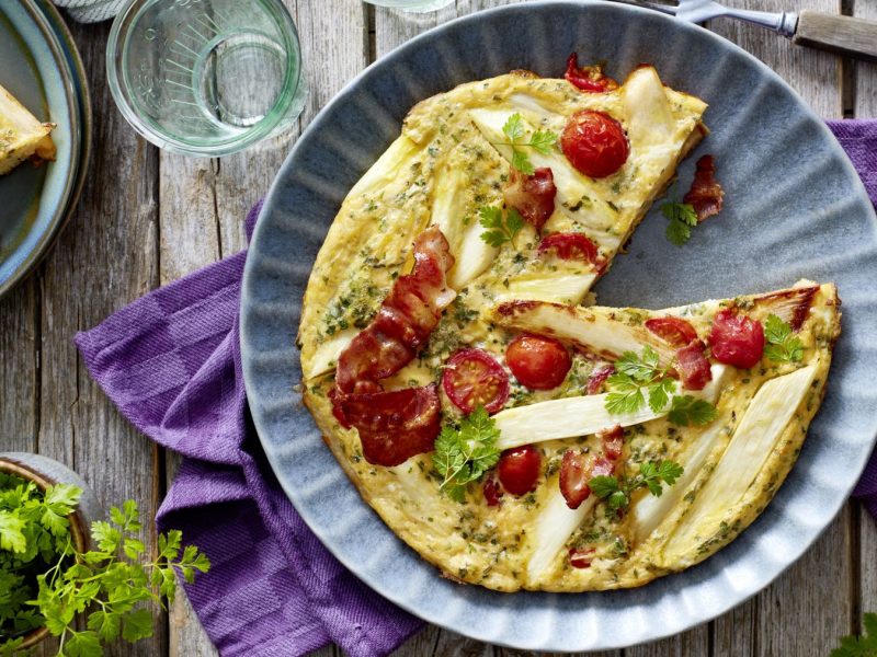 Spargel-Frittata auf einem blauen Teller auf einem lila Geschirrtuch vor hölzernem Hintergrund. Links davon Petersilie und ein Teller mit einem Stück Frittata.