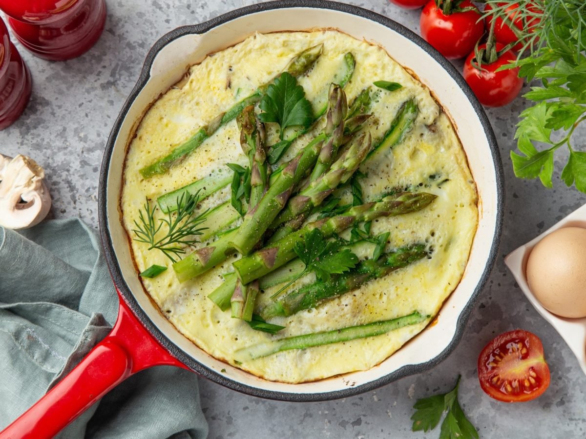 Draufsicht: Ein Spargel-Omelett in einer runden Pfanne. Drumherum Eier in einem Eierkarton, frische Kräuter, Tomaten, Pilze, Salz- und Pfeffermühle: