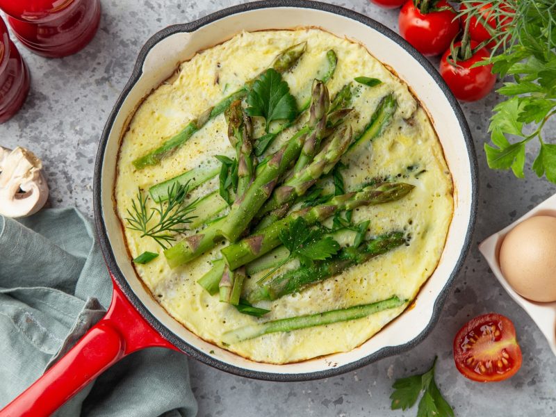 Draufsicht: Ein Spargel-Omelett in einer runden Pfanne. Drumherum Eier in einem Eierkarton, frische Kräuter, Tomaten, Pilze, Salz- und Pfeffermühle: