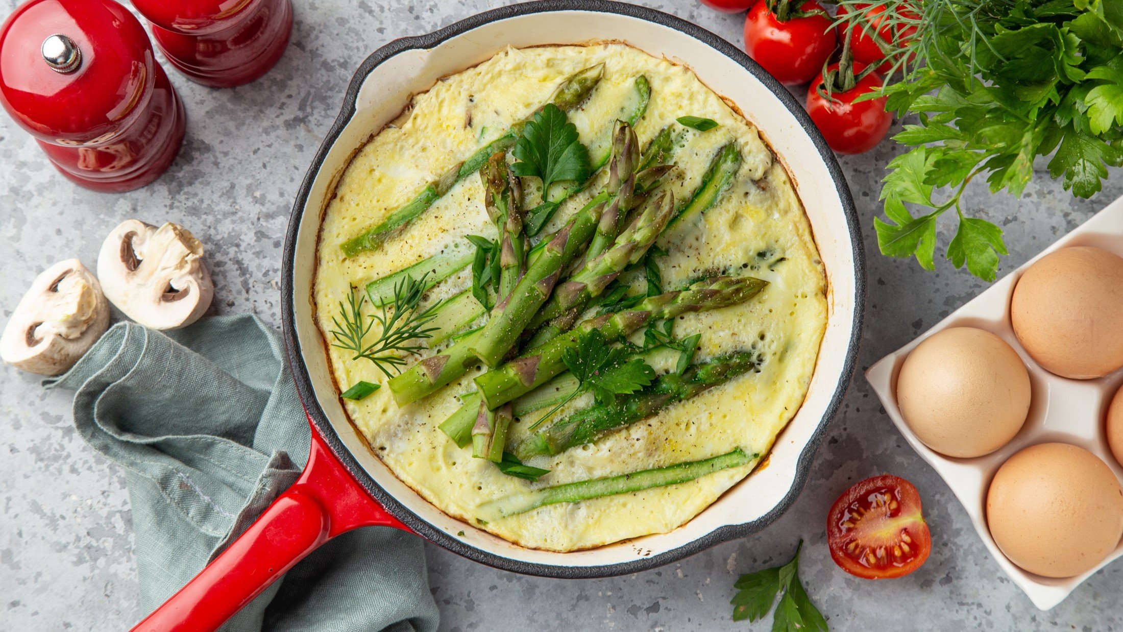 Draufsicht: Ein Spargel-Omelett in einer runden Pfanne. Drumherum Eier in einem Eierkarton, frische Kräuter, Tomaten, Pilze, Salz- und Pfeffermühle: