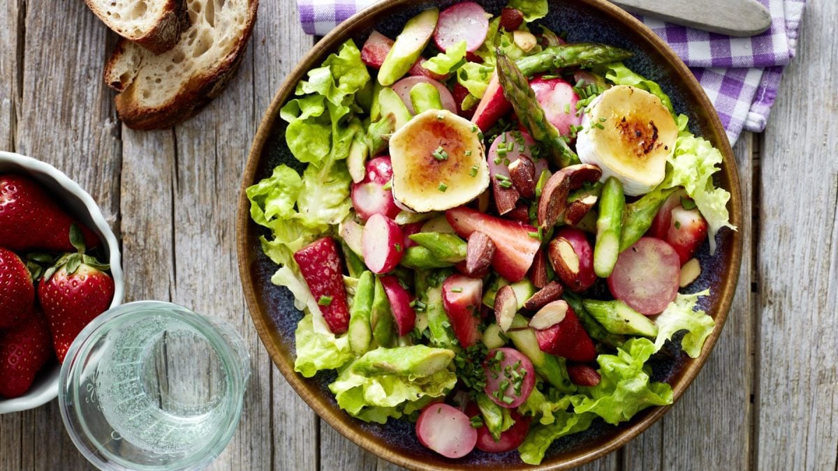 Spargelsalat mit gratiniertem Ziegenkäse in einer braunen Schüssel auf hölzernem Hintergrund. Links Brot, Erdbeeren, ein Glas Wasser und oben auf dem Foto ein blau-weiß-kariertes Geschirrtuch.