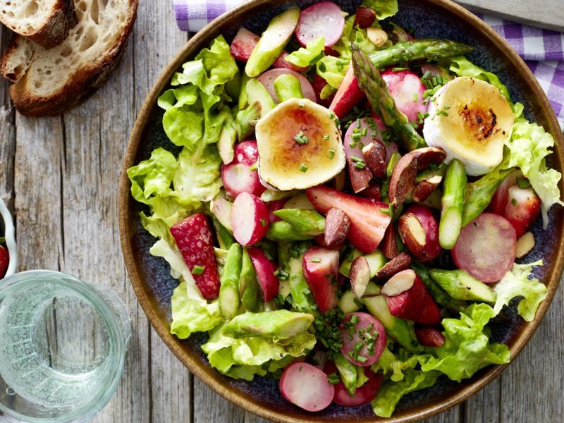 Spargelsalat mit gratiniertem Ziegenkäse in einer braunen Schüssel auf hölzernem Hintergrund. Links Brot, Erdbeeren, ein Glas Wasser und oben auf dem Foto ein blau-weiß-kariertes Geschirrtuch.