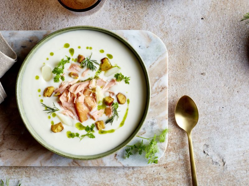 Spargelcreme mit Brezel und Lachs in einer Schüssel auf einem marmoriertem Brett vor beigem Hintergrund. Rechts ein goldener Löffel und links ein beiges Geschirrtuch.
