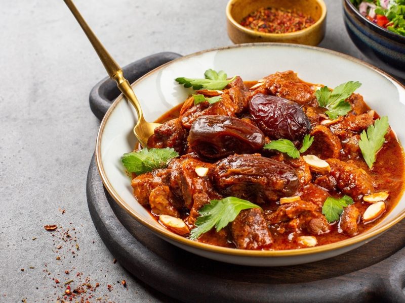 Draufsicht, schräg: eine flache Schüssel mit einer Lamm-Tajine und Dallen und frische Kräutern auf einem runden Teller.