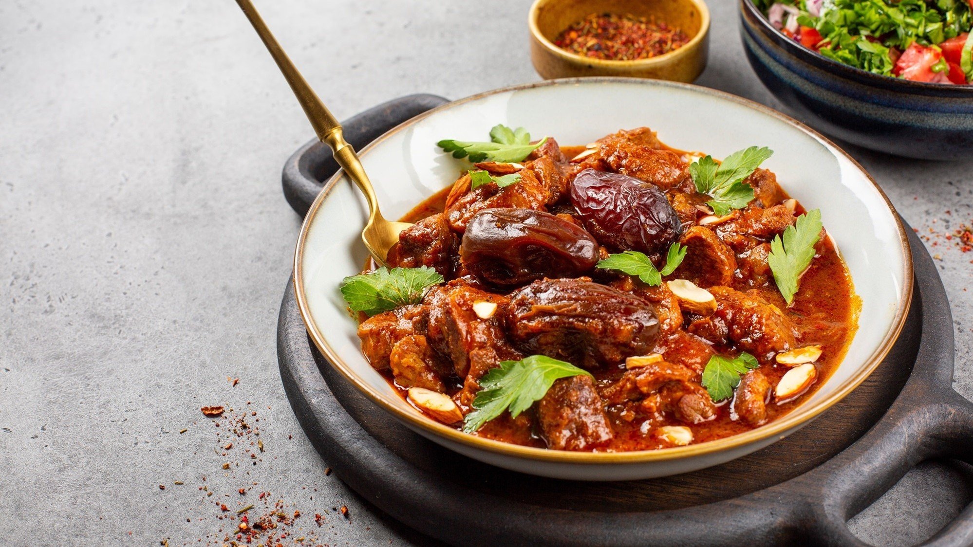 Draufsicht, schräg: eine flache Schüssel mit einer Lamm-Tajine und Dallen und frische Kräutern auf einem runden Teller.