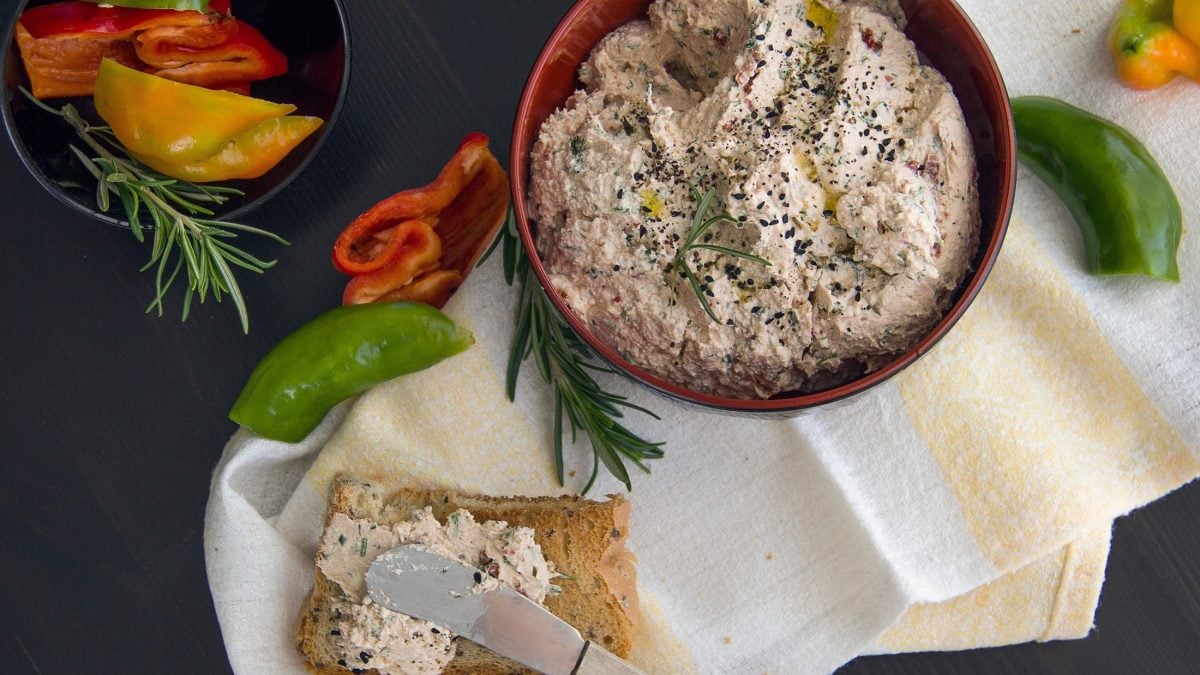 Draufsicht: Dunkler Untergrund, eine runde Schale mit einer Creme. daneben eine Scheibe Brot, die mit der Creme bestrichen ist; Küchenmesser, rohe Paprika-Stücke, frischer Rosmarin.