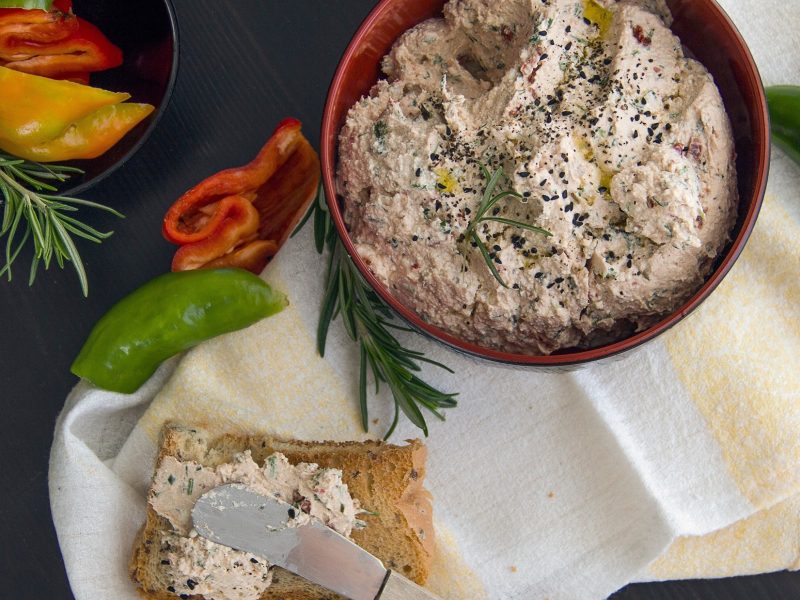 Draufsicht: Dunkler Untergrund, eine runde Schale mit einer Creme. daneben eine Scheibe Brot, die mit der Creme bestrichen ist; Küchenmesser, rohe Paprika-Stücke, frischer Rosmarin.