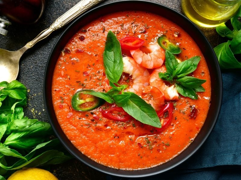 Draufsicht: Eine runde Schüssel mit einer Tomatensuppe mit Garnelen. Drum herum liegen Zitronenscheiben und Basilikumblätter und ein Suppenlöffel.