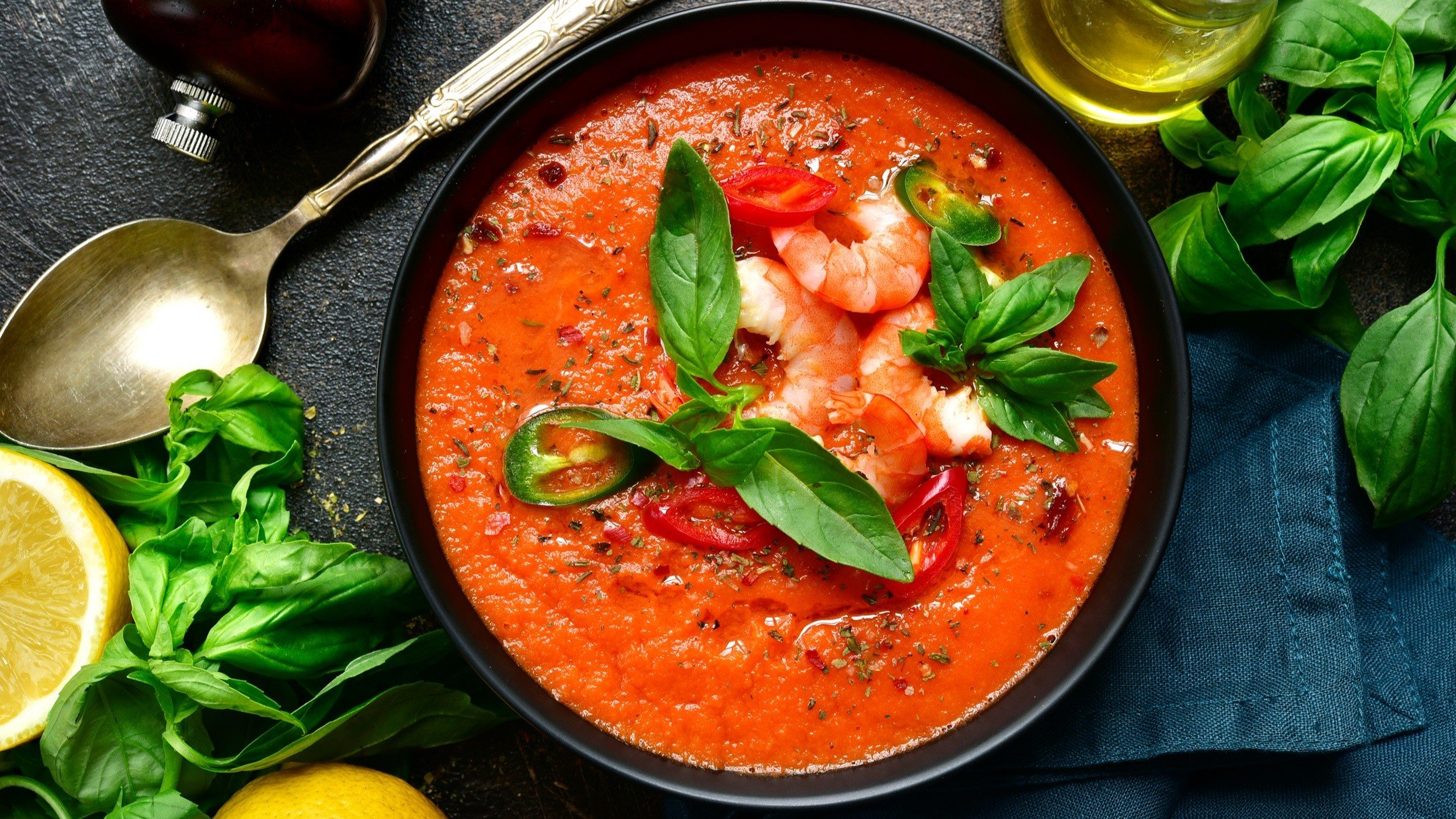 Draufsicht: Eine runde Schüssel mit einer Tomatensuppe mit Garnelen. Drum herum liegen Zitronenscheiben und Basilikumblätter und ein Suppenlöffel.