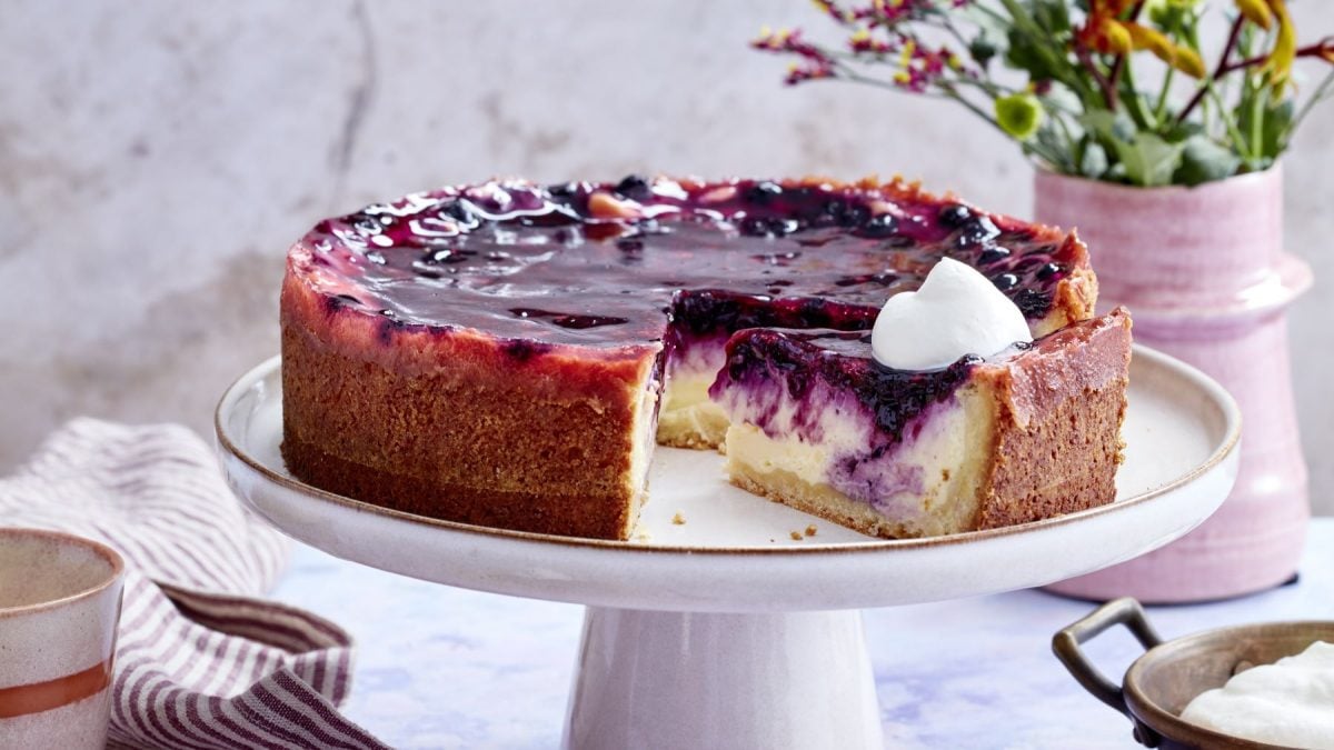 Vanillecreme-Holunder-Torte auf einem weißen Tortenteller vor hellem Hintergrund. Blumen und ein Geschirrtuch sind im Hintergrund zu sehen.