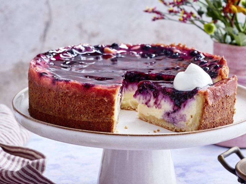 Vanillecreme-Holunder-Torte auf einem weißen Tortenteller vor hellem Hintergrund. Blumen und ein Geschirrtuch sind im Hintergrund zu sehen.
