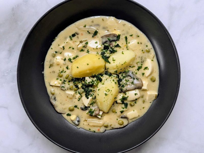 Cremiges Eierragout mit Kartoffeln, Erbsen, Pilzen und Kräutern in heller Sauce auf schwarzem Teller auf Marmorplatte.