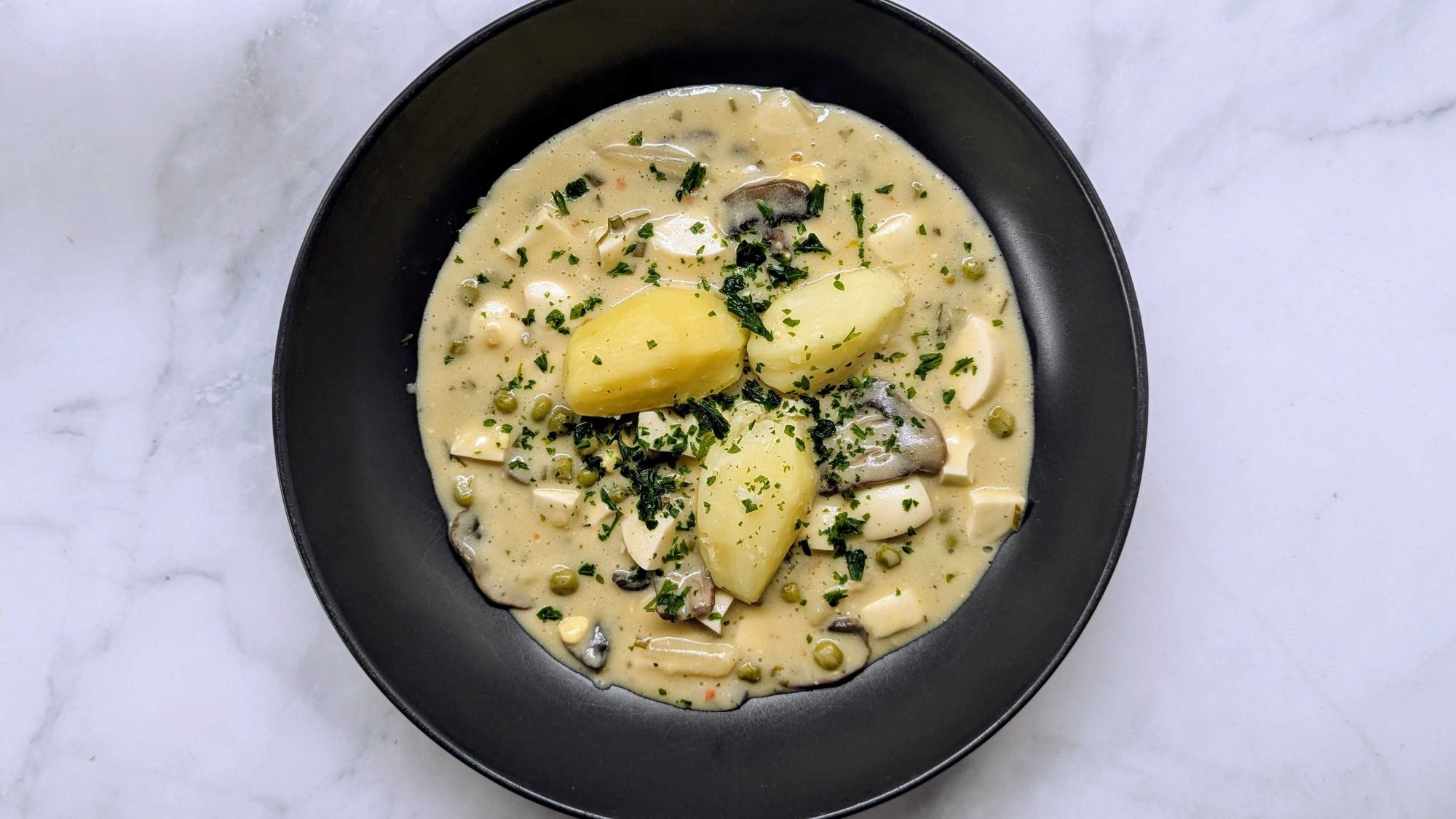 Cremiges Eierragout mit Kartoffeln, Erbsen, Pilzen und Kräutern in heller Sauce auf schwarzem Teller auf Marmorplatte.
