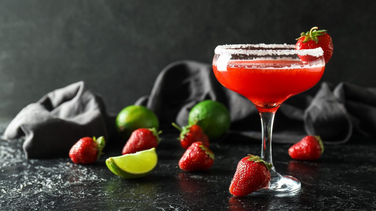 Roter Erdbeer-Daiquiri im Cocktailglas mit Zuckerrand, garniert mit Erdbeere, daneben Limettenstück und frische Erdbeeren.