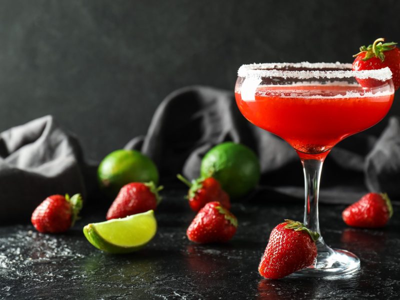 Roter Erdbeer-Daiquiri im Cocktailglas mit Zuckerrand, garniert mit Erdbeere, daneben Limettenstück und frische Erdbeeren.