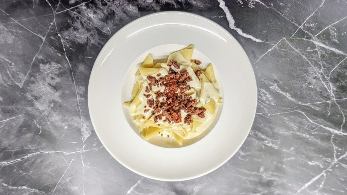 Ein weißer Teller, auf dem Pasta mit Gorgonzola-Sahne-Sauce angerichtet ist. Das Gericht ist belegt mit 'Nduja und gebratener Salsicica.
