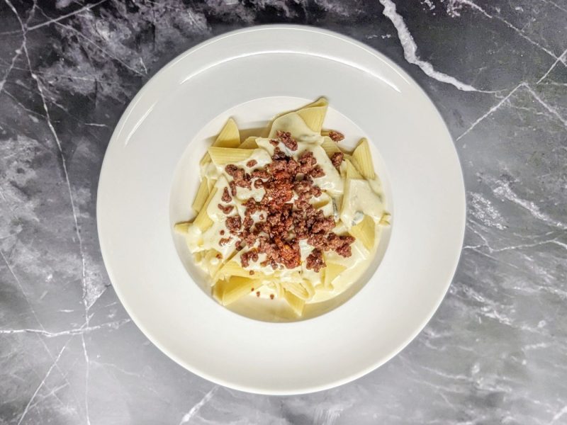 Ein weißer Teller, auf dem Pasta mit Gorgonzola-Sahne-Sauce angerichtet ist. Das Gericht ist belegt mit 'Nduja und gebratener Salsicica.